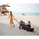 A woman in a brown dress pulls a grey Veer Cruiser XL stroller wagon with three children seated inside along a beach path.