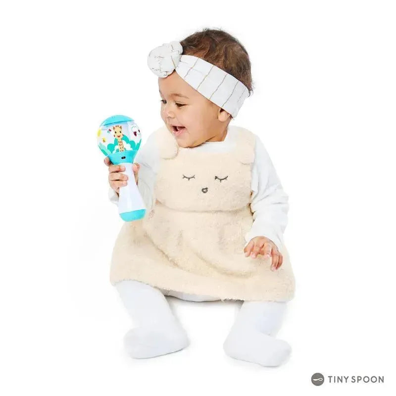 Toddler in a beige plush dress and headband joyfully shakes a colorful toy maraca featuring animals and vibrant patterns.
