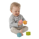A toddler with curly hair smiles while holding a pink and orange rubber ball, with toy cubes nearby.