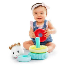 Smiling baby in denim overalls holds a red ring, playing with a colorful stacking toy featuring a plush giraffe.
