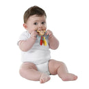 A baby in a white onesie sits and holds a colorful teething ring with a monkey design, looking curiously.