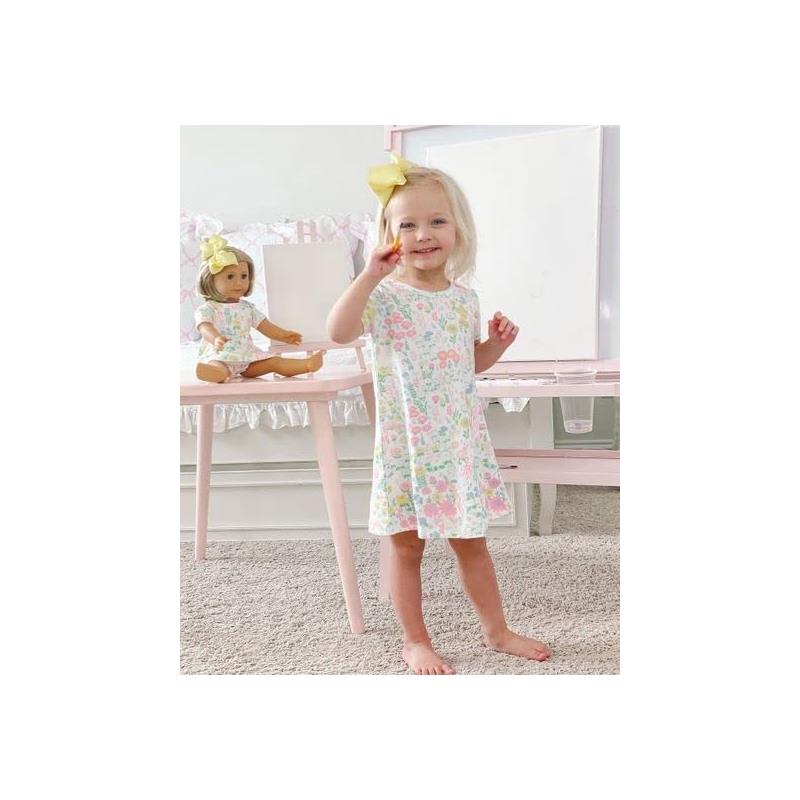 Baby girl in a floral dress smiles while holding a crayon, with a doll in a matching outfit beside her.