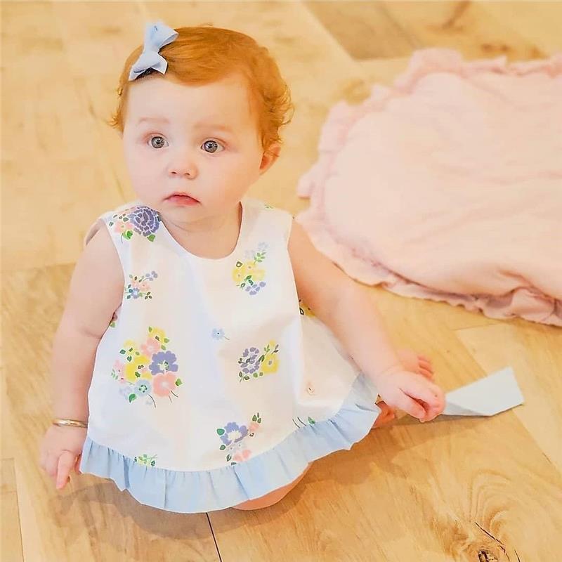 Baby girl with curly red hair and a tiny blue grosgrain hair bow, wearing a floral dress, sitting on a wooden floor.