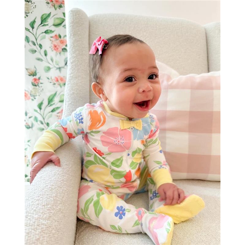 Baby girl smiling while seated in a chair, wearing a floral-patterned onesie and a hot pink hair bow.