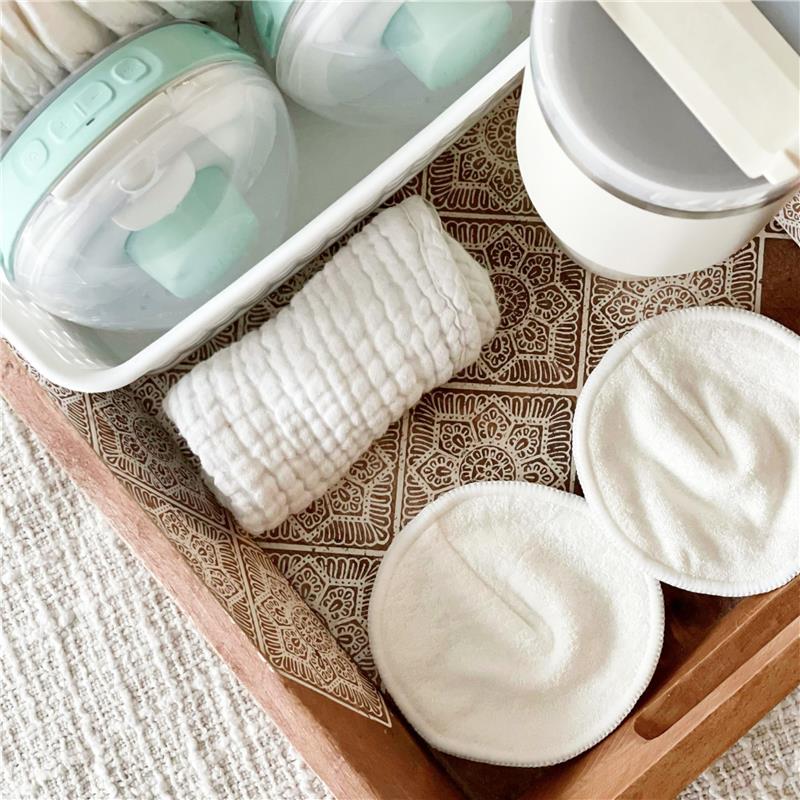 Reusable nursing pads in white, resting on a decorative tray with a rolled cloth and storage containers nearby.