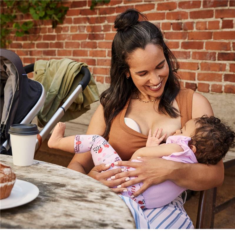 Mother breastfeeding a baby at a rustic table, with a coffee cup and muffin nearby, surrounded by a stroller and greenery.