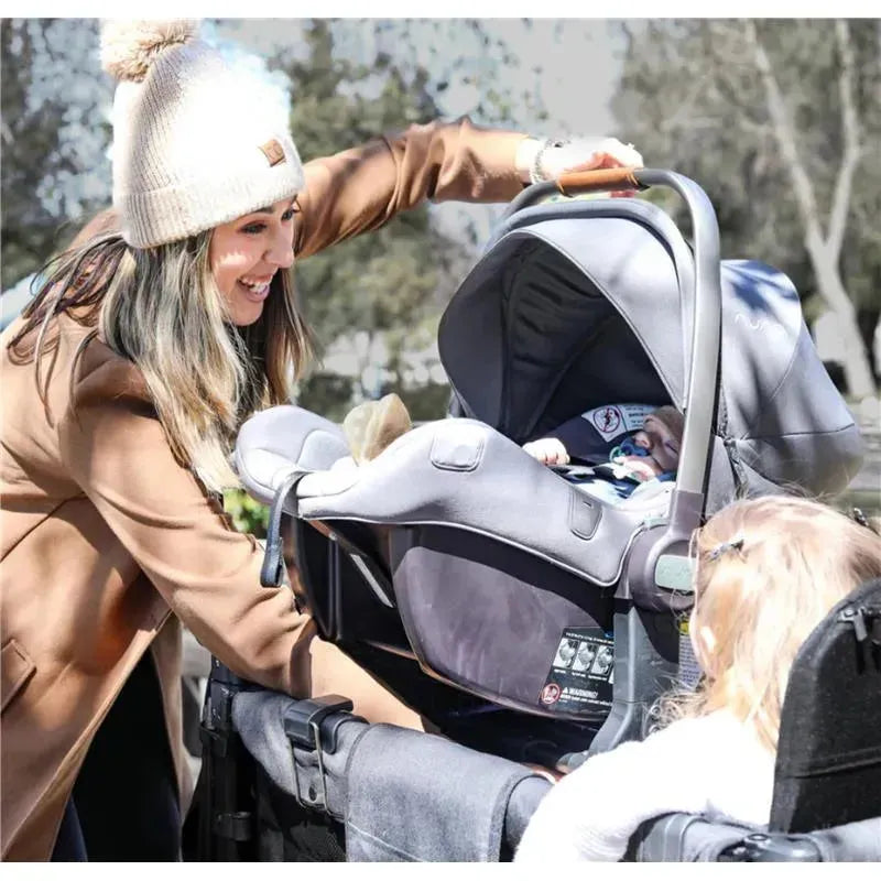 A woman in a beige coat is placing an infant car seat into a stroller while a toddler sits nearby, wearing sunglasses.