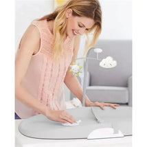 A woman wipes a grey Skip Hop wipe-clean changing pad, which features a toy mobile with a sun and cloud, in a nursery setting.
