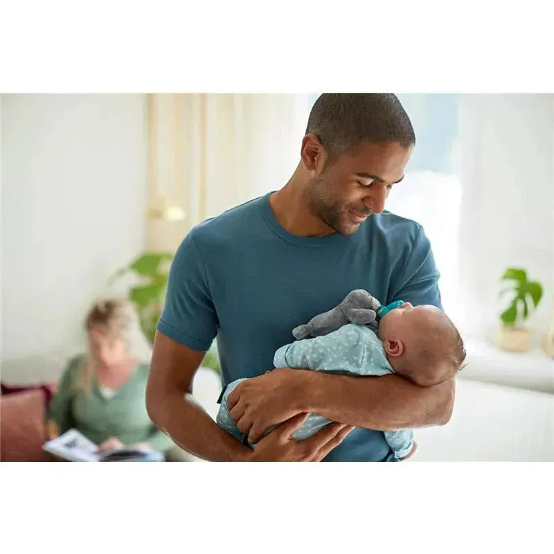 A father gently holds his baby, who is snuggling a soft gray seal toy, both appearing content in a bright room.